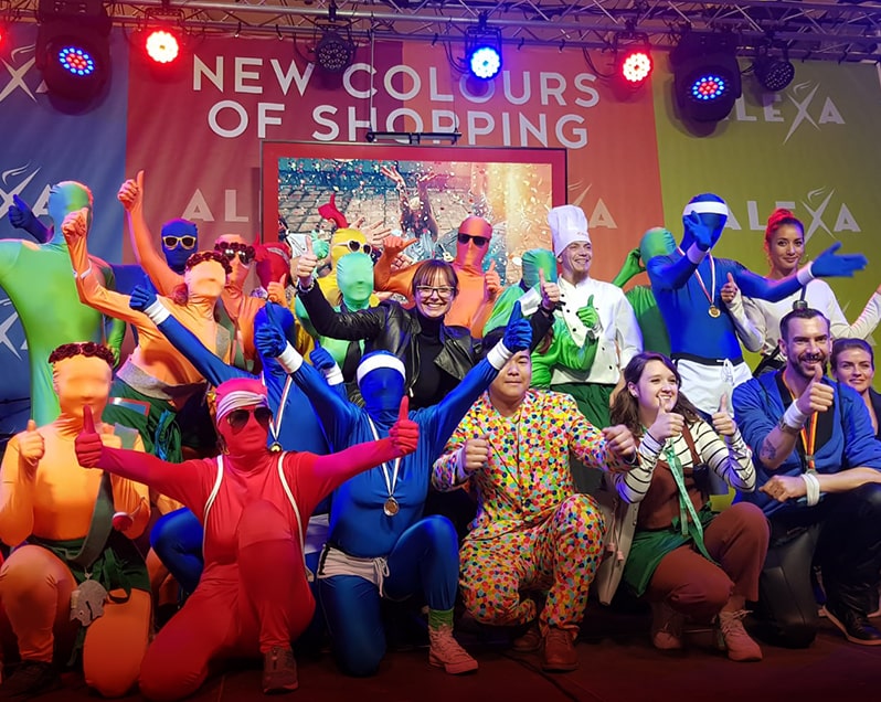 Mehrere Menschen aus dem Team des Events ALEXA NEW COLOURS OF SHOPPING sind in drei Reihen hintereinander aufgestellt. Einige von ihnen tragen bunte Morph-Kostüme, die den gesamten Körper bedecken. Alle Menschen sind ausgelassen und feiern den Abschluss des Events im Metropolit Court des ALEXA Shoppingcenters. Im Hintergrund ist ein Teil der Bühne sichtbar, mit dem Logo des Events.
