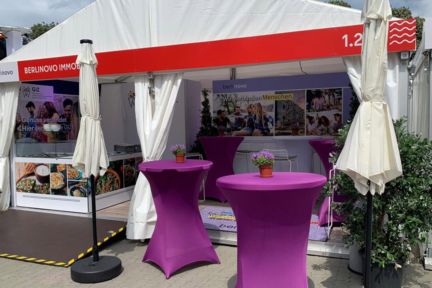 Ein Stand von berlinovo auf dem Hoffest des Regierenden Bürgermeisters von Berlin, mit einem Zelt, Stehtischen in violettfarbenen Hussen, großen, weissen Schirmen, einem Counter für Zubereitung und Ausgabe von orientalischen Snacks, zwei Präsentationswänden und Pflanzen als Dekoration.