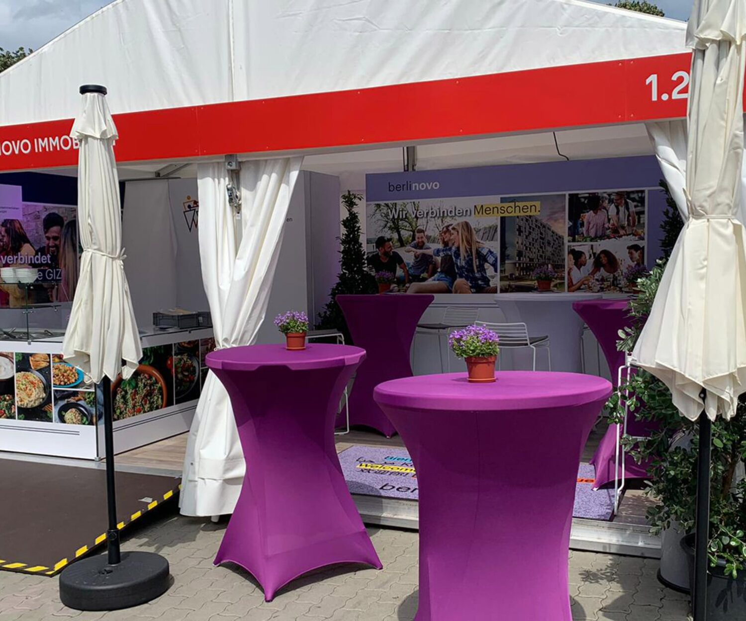 Ein Stand von berlinovo auf dem Hoffest des Regierenden Bürgermeisters von Berlin, mit einem Zelt, Stehtischen in violettfarbenen Hussen, großen, weissen Schirmen, einem Counter für Zubereitung und Ausgabe von orientalischen Snacks, zwei Präsentationswänden und Pflanzen als Dekoration.