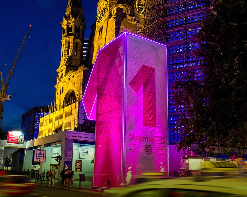 Eine Nachtaufnahme auf dem Breitscheidplatz in Berlin, auf dem das Event Berlin Celebrates IFA stattfindet. Eine riesige, in Magentafarbe leuchtende Konstruktion in Form der Zahl 1 steht im Vordergrund. Im Hintergrund ist ein Ausschnitt der Gedächtniskirche zu sehen. Trendshow Kurfürstendamm City West