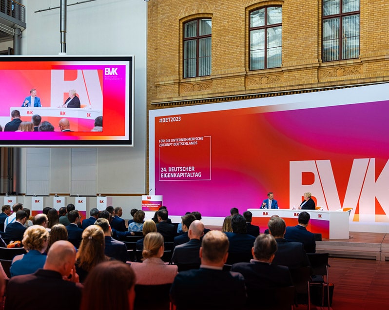 Im Vordergrund sind mehrere Zuschauerreihen zu sehen. Die Zuschauer nehmen am 24. Deutschen Eigenkapitaltag teil. Im Hintergrund, rechts auf dem Podium sprechen prominente Referenten miteinander. Links auf dem Podium ist eine große Leinwand zu sehen, auf die das Gespräch projiziert wird. Hinter dem Podium befindet sich eine große Bühnenwand mit BVK-Branding.