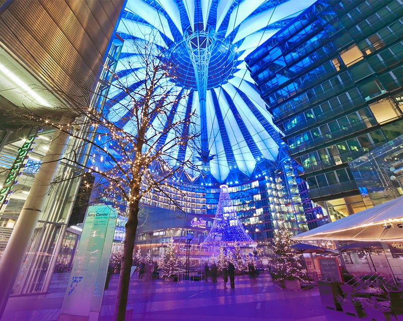 Innenhof des Sony Centers in Berlin, links ein Baum mit Weihnachtsbeleuchtung, dahinter einige Tannenbäume mit Lichterketten, oben das blau leuchtende Dach des Sony Centers, im Hintergrund ein weihnachtlicher Trichter aus von oben herabhängender, blau leuchtender Lichterketten. SONY Center am Potsdamer Platz Weihnachtsaktionen Magic Christmas