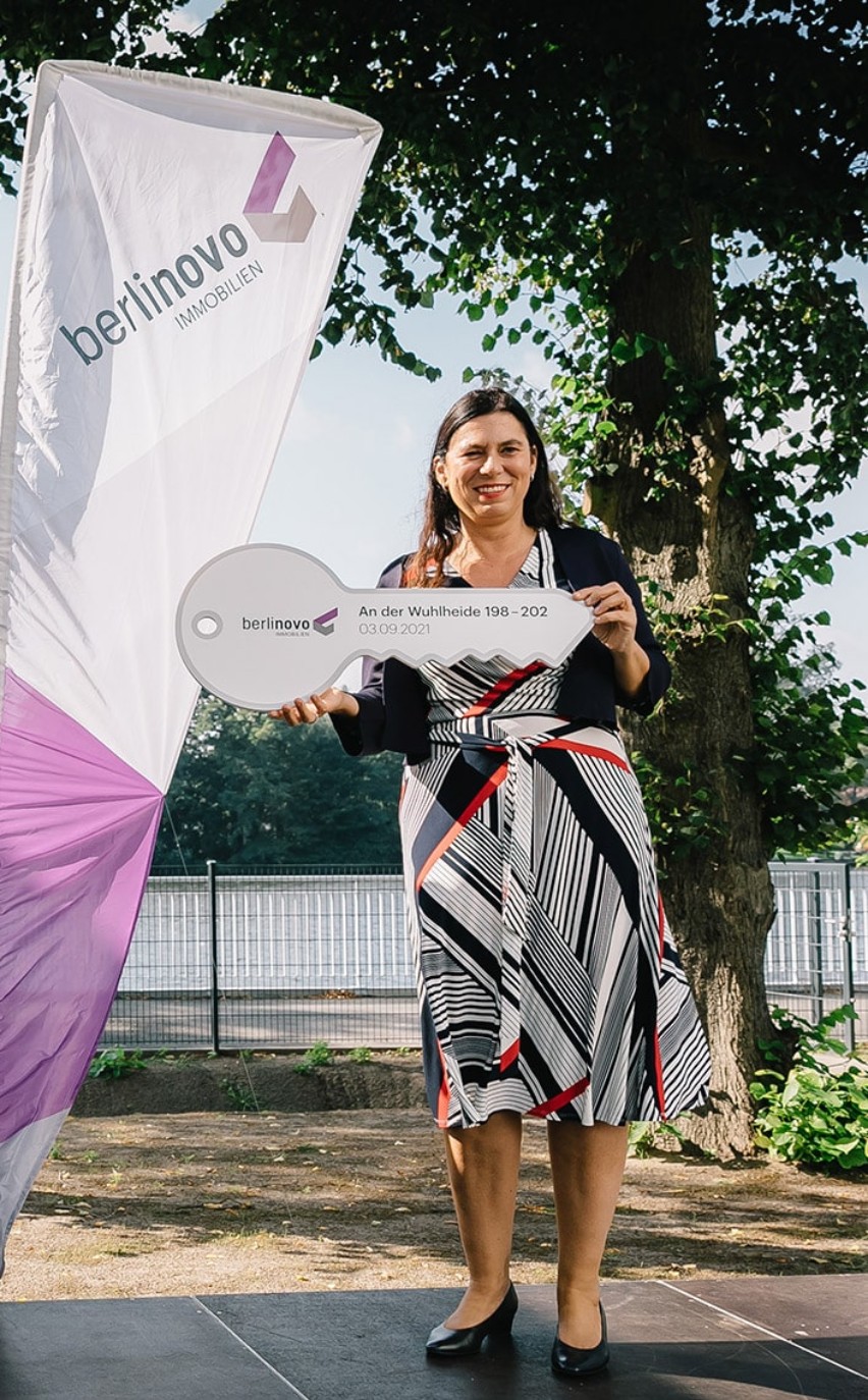 Eine Person auf der Bühne bei der Schlüsselübergabe eines Bauprojekts von berlinovo in der Wuhlheide. Sie präsentiert einen symbolischen Schlüssel, der aus Anlass der Schlüsselübergabe angefertigt wurde. Hinter den Personen sind Banner mit Branding von berlinovo.