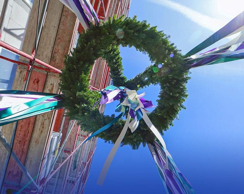 Auf dem Richtfest eines Bauprojekts von berlinovo in der Wuhlheide wird ein Richtkranz mit weissen, grünen und violetten Bändern, von unten Fotografiert, entlang eines Baugerüsts nach oben gezogen.