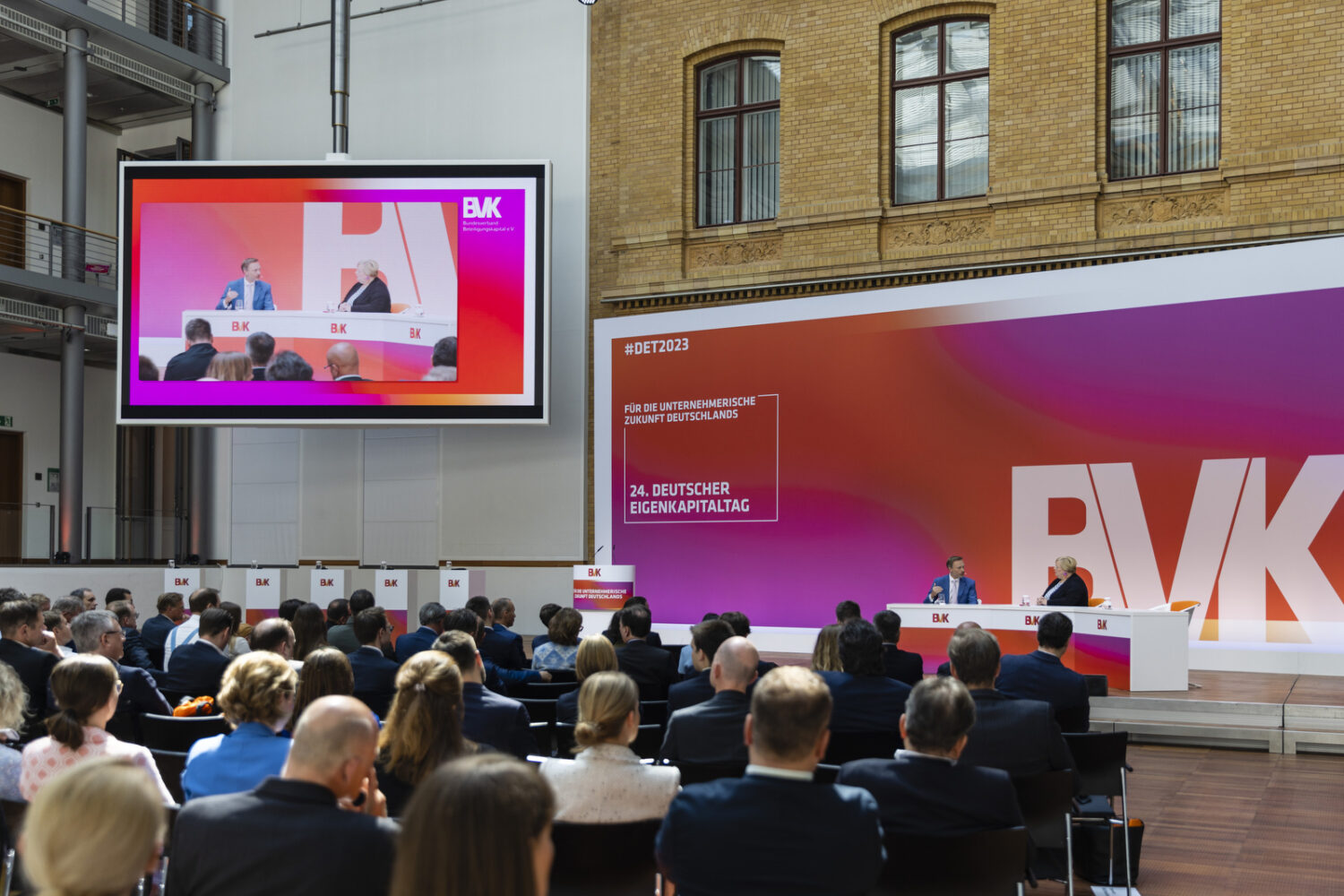 Im Vordergrund sind mehrere Zuschauerreihen zu sehen. Die Zuschauer nehmen am 24. Deutschen Eigenkapitaltag teil. Im Hintergrund, rechts auf dem Podium sprechen prominente Referenten miteinander. Links auf dem Podium ist eine große Leinwand zu sehen, auf die das Gespräch projiziert wird. Hinter dem Podium befindet sich eine große Bühnenwand mit BVK-Branding.