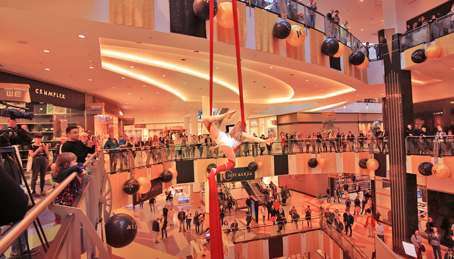 Im Innenraum des ALEXA Shoppingcenters in Berlin findet das Event 10 JAHRE ALEXA statt. Eine Frau mit langem, blondem Haar, gekleidet im Stil der Goldenen Zwanziger Jahre, zeigt eine Luftakrobatik-Show. Sie schwingt durch die Luft mithilfe eines langen roten Stoffbandes, das an der Decke des Innenraums befestigt sind. Viele Besucher des ALEXA Berlin, die sich auf den Etagen des Innenraums befinden, beobachten die Show.