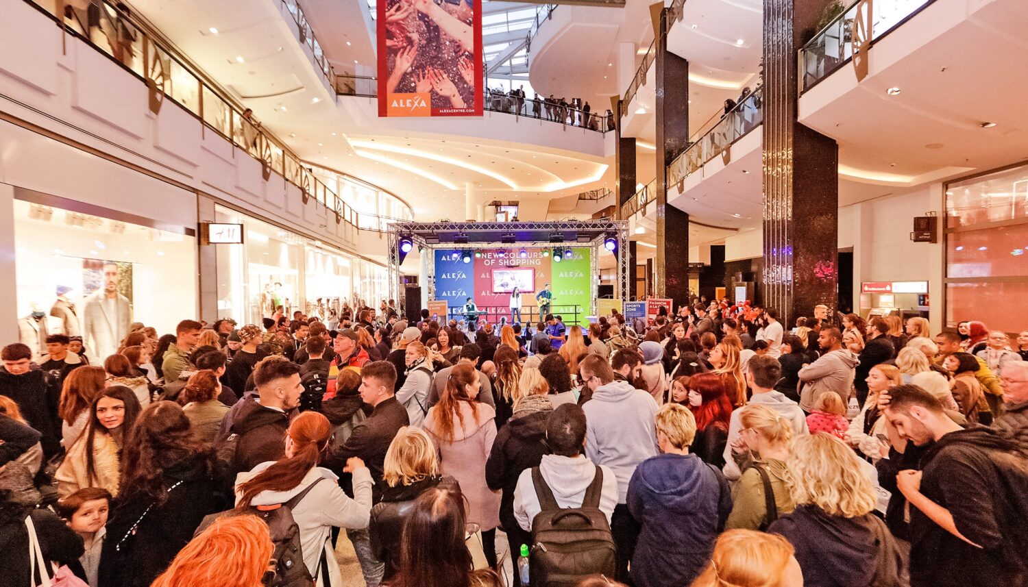 Im Metropolis Court des ALEXA Shoppingcenters findet eine Musikshow im Rahmen des Events ALEXA NEW COLOURS OF SHOPPING. Im Vordergrund sind zahlreiche Zuschauer zu sehen, die eine Musikshow beim verfolgen. Im Hintergrund steht die Bühne mit einer dreiköpfigen Musikband.