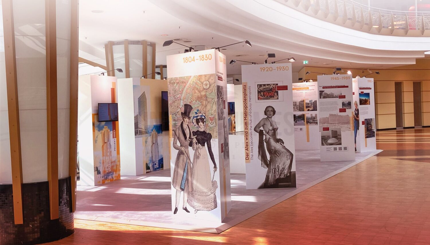 Im Innenraum des ALEXA Shoppingcenters findet die Ausstellung ZUKUNFT ALEXANDERPLATZ. EIN PLATZ im WANDEL statt. Zu sehen sind dreieckige Stellwände mit historischen Figuren und Fotos vom Alexanderplatz, dahinter rechteckige Stellwände mit integrierten Bildschirmen, auf welchen Videos über die Gegenwart und Zukunft vom Alexanderplatz informiert wird.