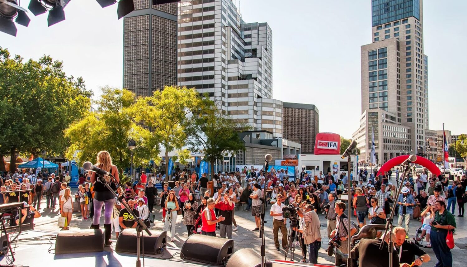 Eine Aufnahme auf dem Breitscheidplatz in Berlin, auf dem das Event Berlin Celebrates IFA stattfindet. Im Vordergrund ist eine Bühne zu sehen. Ein Musikerin spielt vor zahlreichem Publikum, einem Kameramann und mehreren Fotografen. Im Hintergrund sind Hochhäuser zu sehen.