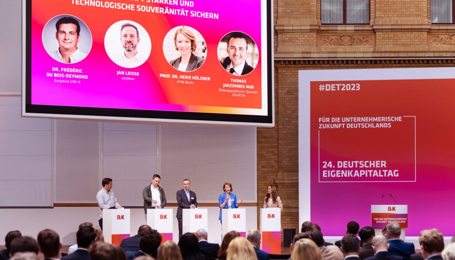 Auf dem Podium des 24. Deutschen Eigenkapitaltags führen fünf Referenten, hinter Rednerpulten mit BVK-Branding stehend, eine Podiumsdiskussion durch. Hinter den Referenten und rechts daneben ist eine große Videoleinwand und eine große Bühnenwand mit BVK-Branding zu sehen. Vor den Referenten sind mehrere Zuschauerreihen zu sehen.