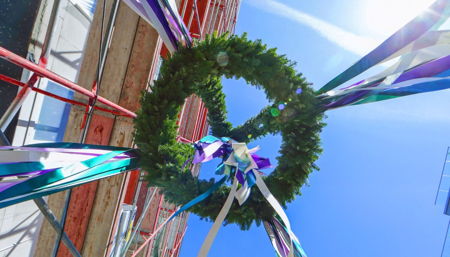 Auf dem Richtfest eines Bauprojekts von berlinovo in der Wuhlheide wird ein Richtkranz mit weissen, grünen und violetten Bändern, von unten Fotografiert, entlang eines Baugerüsts nach oben gezogen.