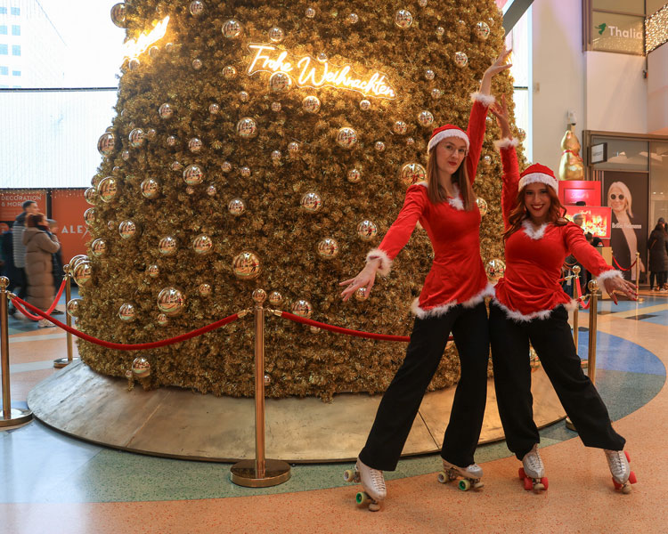 Zwei Frauen in roten Weihnachtskostümen und Rollschuhen posieren vor einem riesigen, gold‑verzierten Weihnachtsbaum in der ALEXA‑Mall in Berlin.