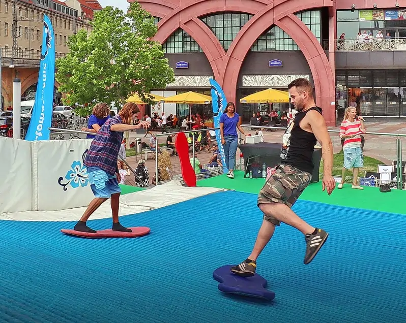 Für das Event ALEXA – THE WAVE wurde vor dem Eingang zum ALEXA Shoppingcenter eine künstliche, endlose Surfing-Welle aufgebaut. Im Vordergrund sind zwei junge Männer zu sehen, die auf Surfbrettern die endlose Welle reiten. Im Hintergrund ist die Moderatorin des Events und wartende Besucher auf dem Ravelinplatz vor dem Eingang zum ALEXA Berlin zu sehen.