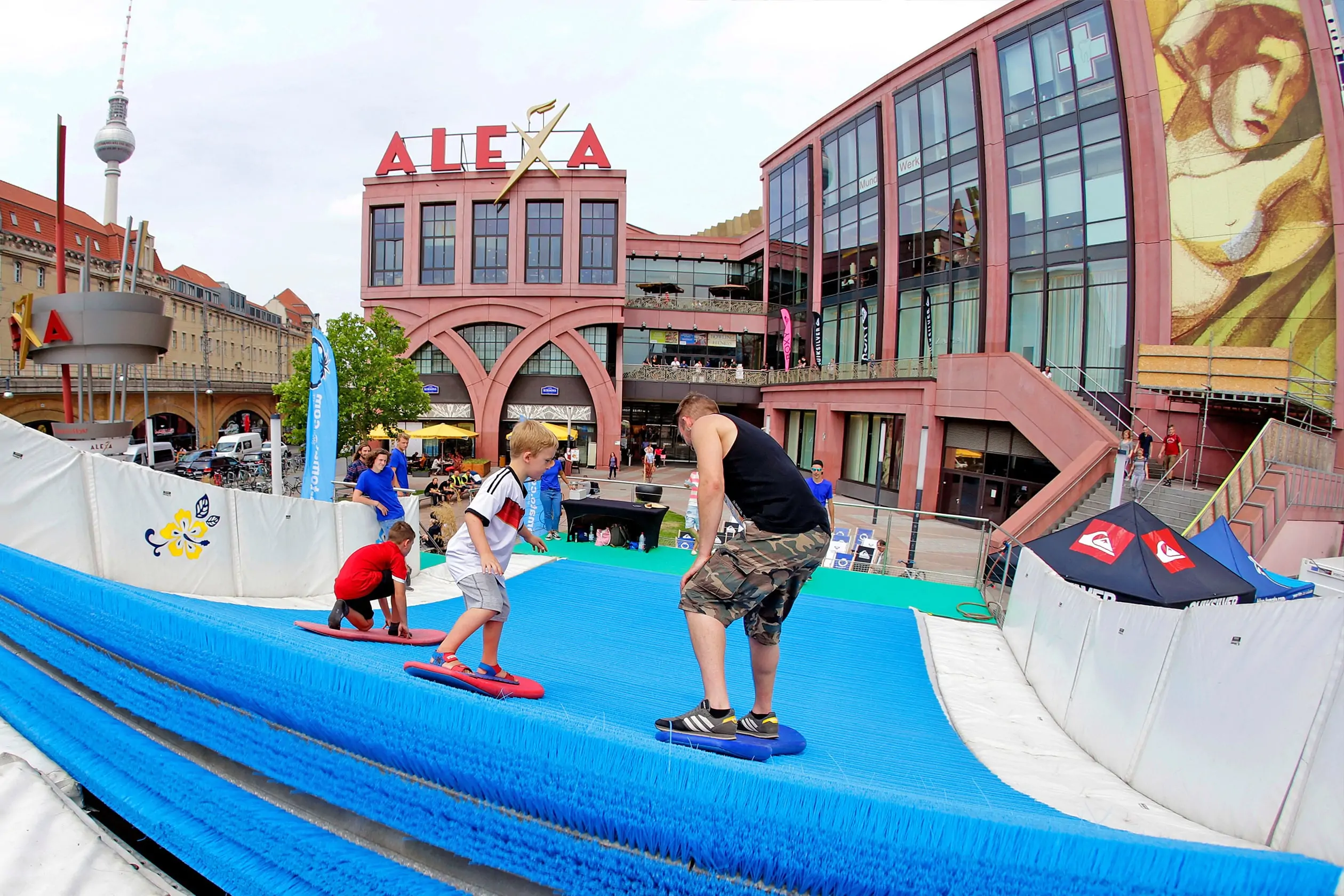 Für das Event ALEXA – THE WAVE wurde vor dem Eingang zum ALEXA Shoppingcenter eine künstliche, endlose, blaue Surfing-Welle aufgebaut. Im Vordergrund ist ein junger Mann und ein Kind zu sehen, die auf Surfbrettern die endlose Welle reiten. Im Hintergrund ist der Ravelinplatz vor dem Eingang zum ALEXA Berlin zu sehen.
