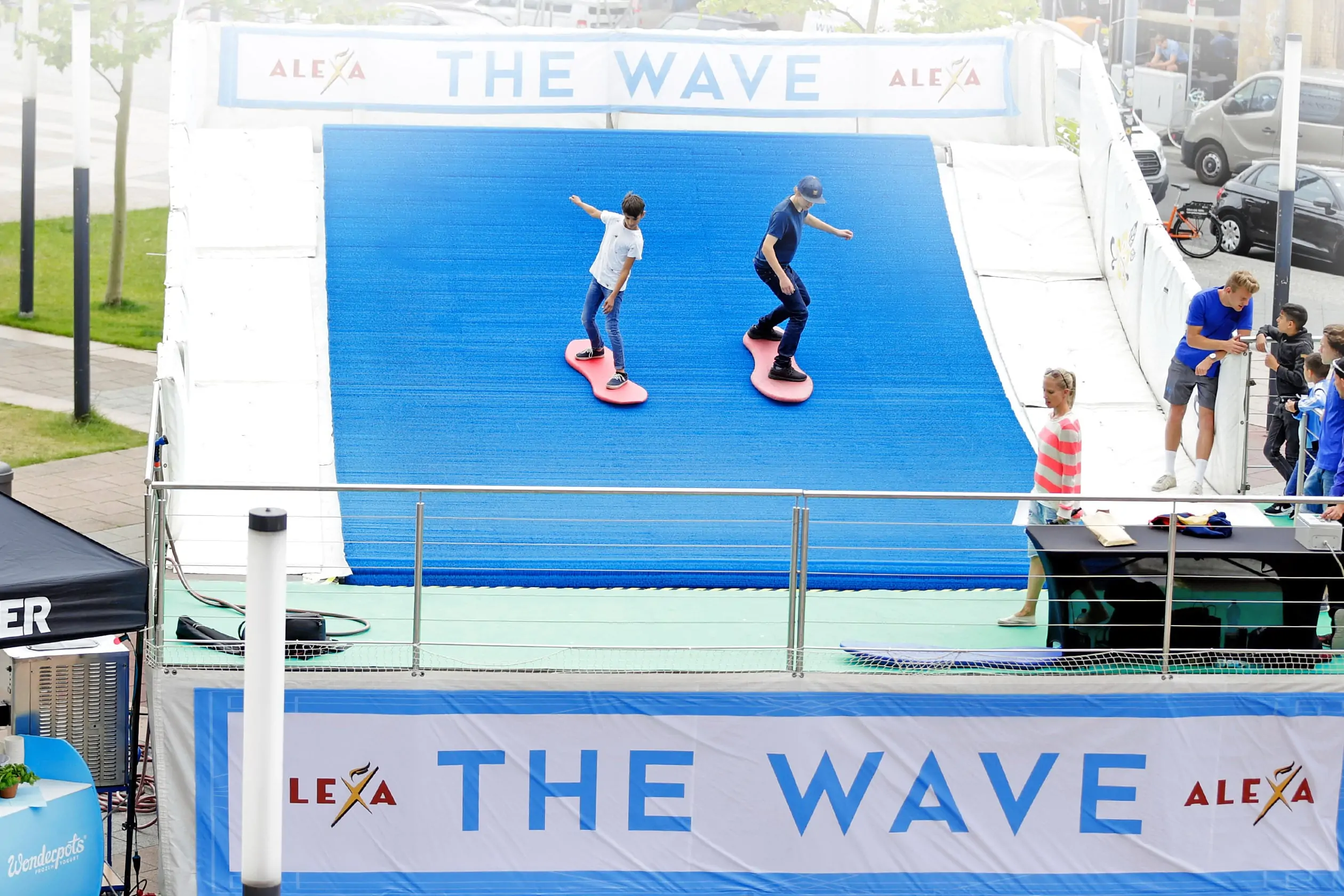 Vor dem Eingang zum ALEXA Shoppingcenter in Berlin wurde eine künstliche, endlose Surfing-Welle aufgebaut. Im Vordergrund sind zwei junge Männer zu sehen, die auf Surfbrettern die künstliche, blaue, endlose Welle reiten. Im Hintergrund und unterhalb der Wellenanlage erscheinen Banner mit dem Schriftzug THE WAVE und Logos von ALEXA.