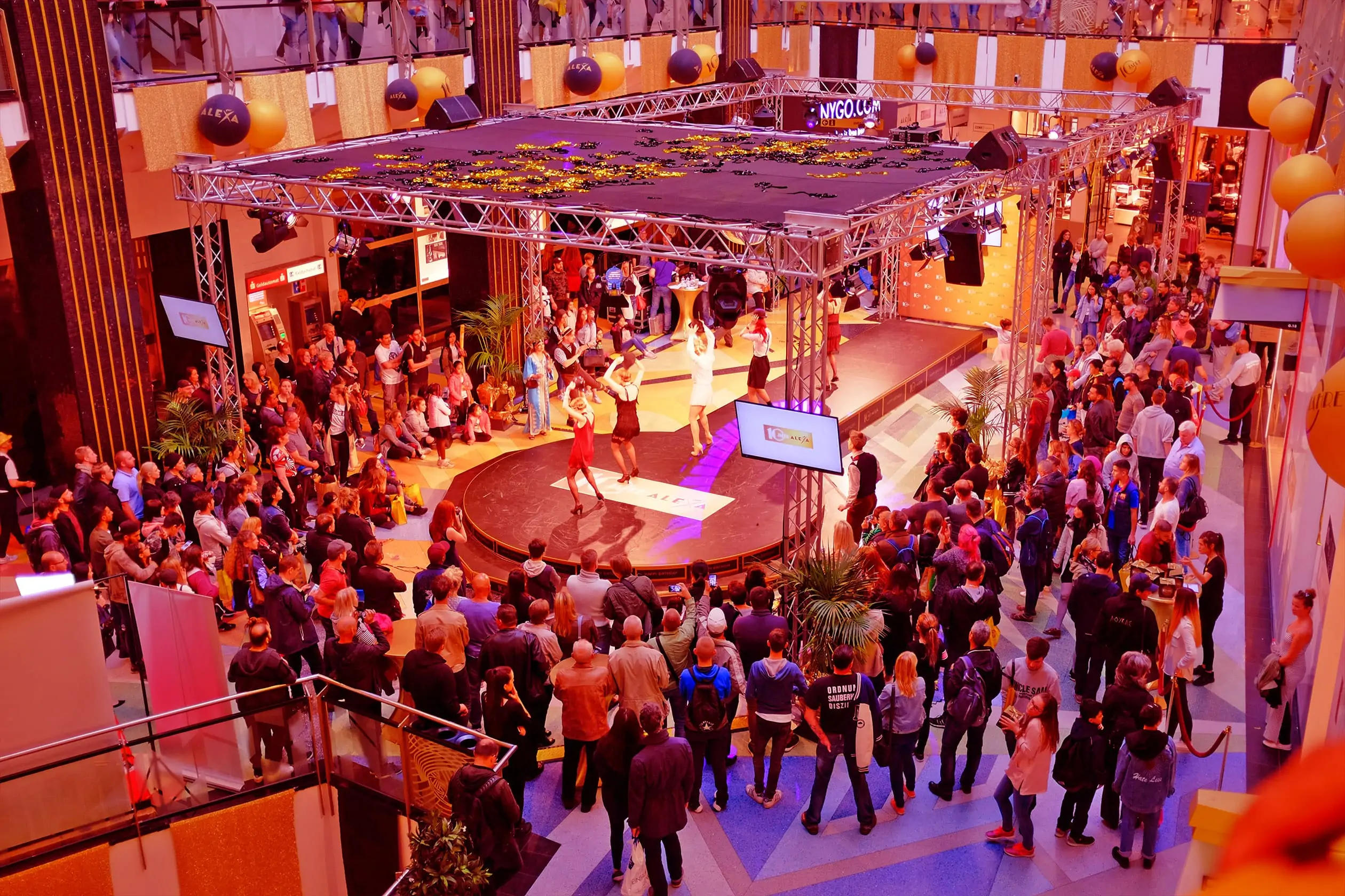 Eine Tanzshow findet auf dem Metropolis Court im ALEXA Shoppingcenter statt. Mehrere Tänzer*innen, gekleidet im Stil der Goldenen Zwanziger Jahre führen Tänze aus den „Roaring Twenties“ vor. Der Metropolis Court ist mit einer Dekoration und dem Logo 10 Jahre ALEXA ausgeschmückt. Viele Besucher beobachten die Show.