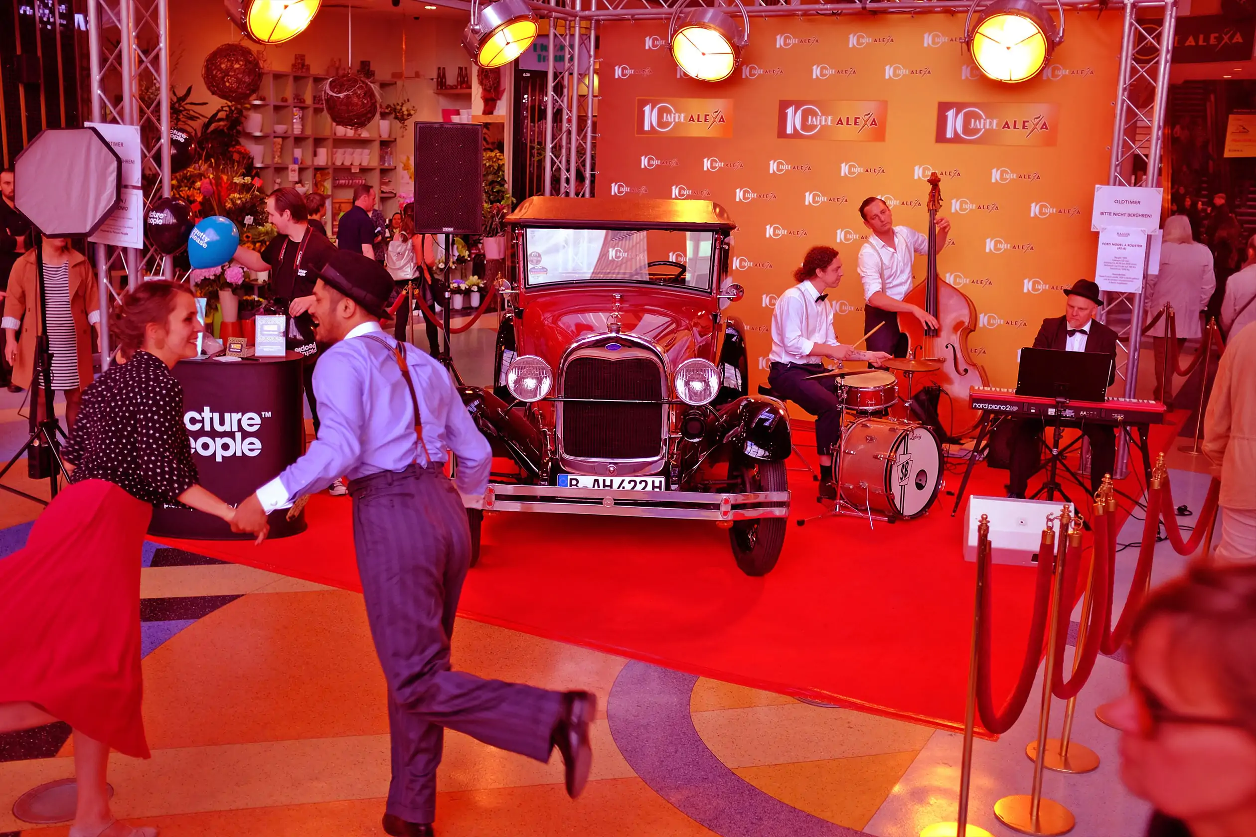 Ein Mann und eine Frau, gekleidet im Stil der Goldenen Zwanziger Jahre, zeigen eine Tanz-Show im Innenraum des ALEXA Shoppingcenter in Berlin. Im Hintergrund spielt eine dreiköpfige Band: Ein Keybord-Spieler, ein Kontrabass-Spieler und ein Schlagzeuger auf einem roten Teppich. Links neben der Band ist ein Oldtimer aus den Goldenen Zwanzigern zu sehen. Die Bühne ist mit dem Logo 10 Jahre ALEXA ausgeschmückt.