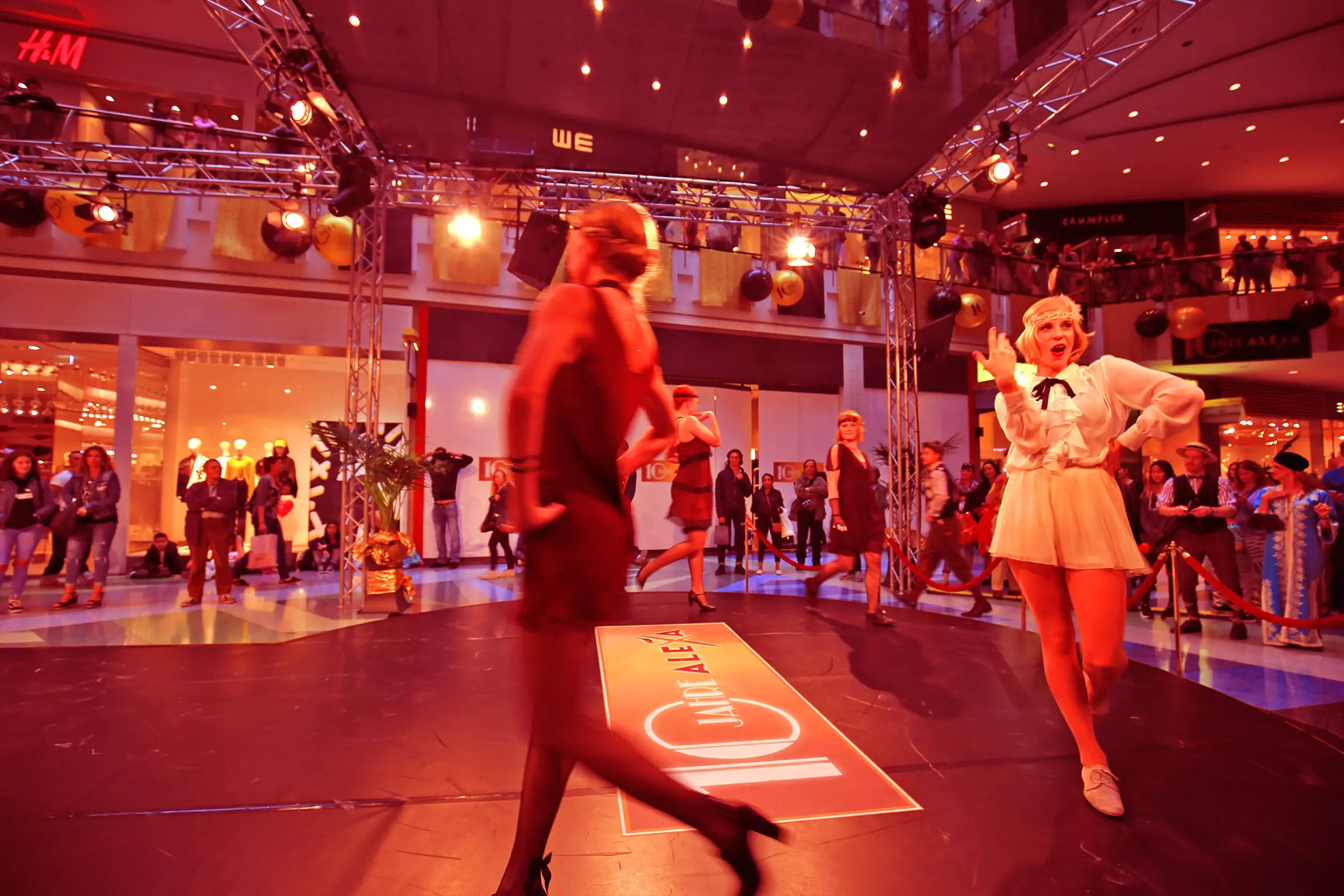 Eine Tanz- und Modeshow im Stil der Goldenen Zwanziger Jahre findet auf dem Metropolis Court im ALEXA Shoppingcenter statt. Mehrere Tänzerinnen führen Tänze und Kostüme aus den „Roaring Twenties“ vor. Die Tanzbühne des Metropolis Court ist mit dem Logo 10 Jahre ALEXA ausgeschmückt. Viele Besucher beobachten die Show.