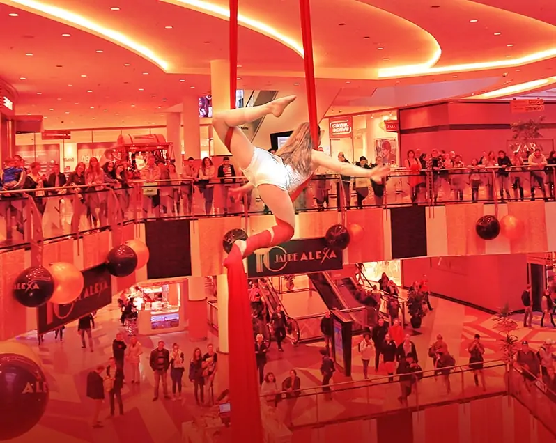 Eine Frau mit langem, blondem Haar, gekleidet im Stil der Goldenen Zwanziger Jahre, zeigt eine Luftakrobatik-Show im Innenraum des ALEXA Shoppingcenter in Berlin, im Rahmen des Events 10 Jahre ALEXA. Sie schwingt durch die Luft mithilfe eines vertikalen, langen, roten Stoffbandes, das an der Decke des Innenraums befestigt sind. Viele Besucher des ALEXA Berlin, die sich auf den Etagen des Innenraums befinden, beobachten die Show.