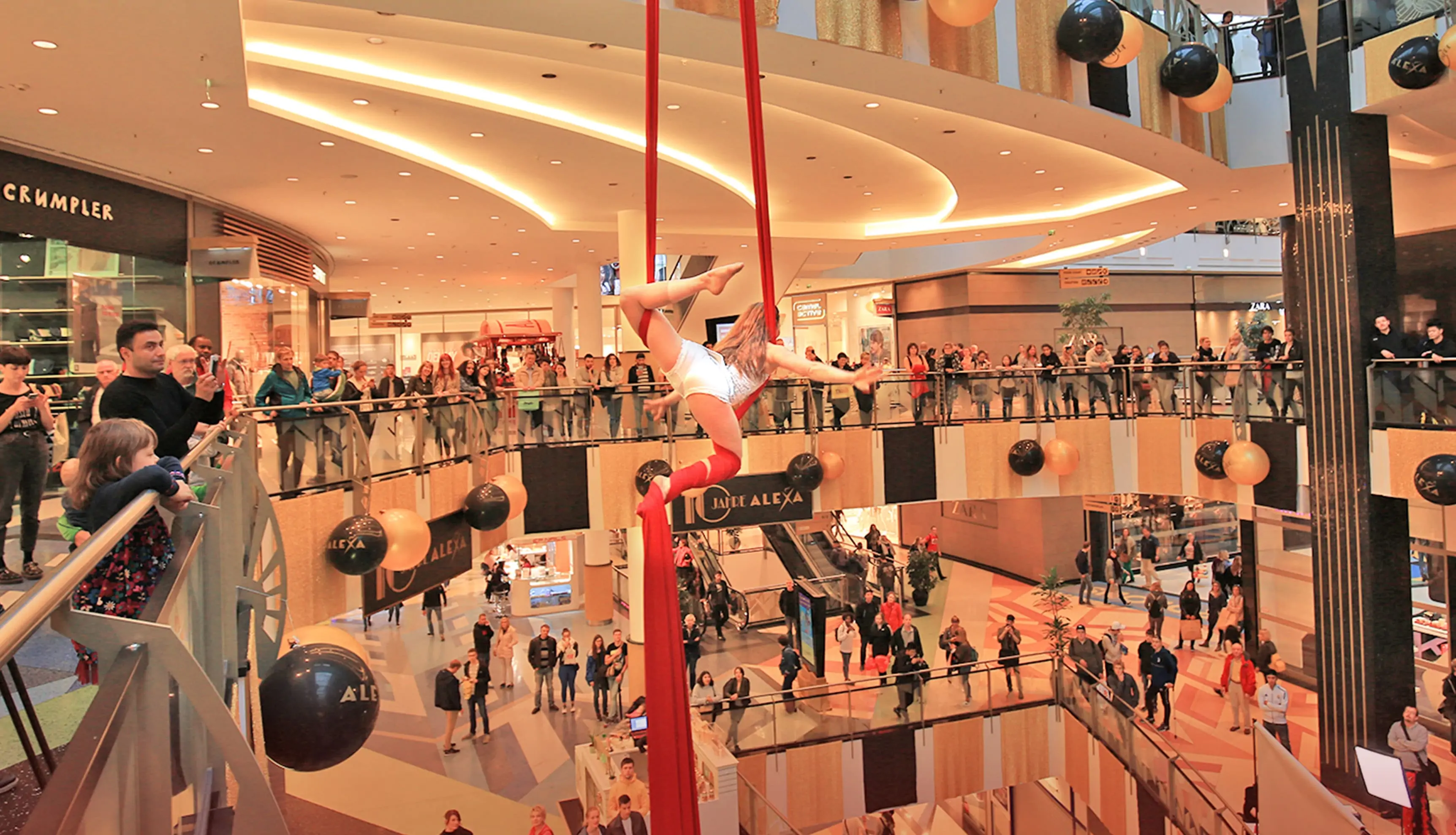 Eine Frau mit langem, blondem Haar, gekleidet im Stil der Goldenen Zwanziger Jahre, zeigt eine Luftakrobatik-Show im Innenraum des ALEXA Shoppingcenter in Berlin, im Rahmen des Events 10 Jahre ALEXA. Sie schwingt durch die Luft mithilfe eines vertikalen, langen, roten Stoffbandes, das an der Decke des Innenraums befestigt sind. Viele Besucher des ALEXA Berlin, die sich auf den Etagen des Innenraums befinden, beobachten die Show.