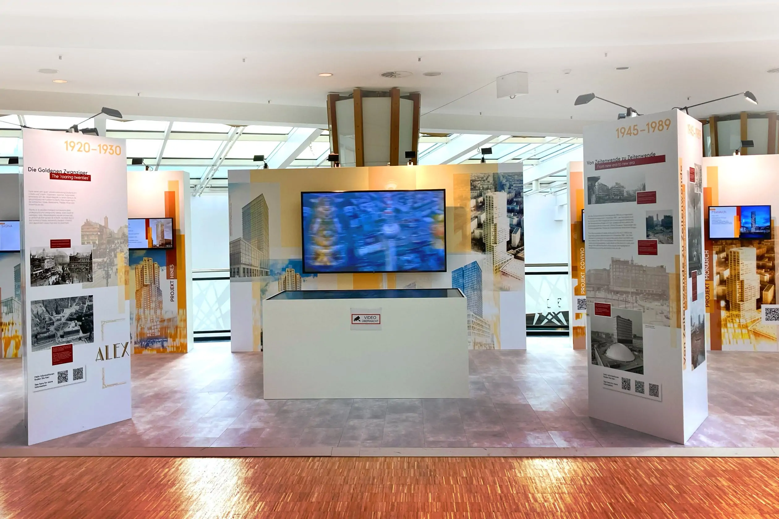 Frontale Ansicht der Informationswand der Ausstellung „ZUKUNFT ALEXANDERPLATZ. EIN PLATZ im WANDEL.“ im der 3. Etage des ALEXA Shoppingcenters. Auf der Informationswand ist ein Bildschirm befestigt, auf dem ein Video über die Vergangenheit, Gegenwart und Zukunft vom Alexanderplatz abläuft. Vor der Informationswand steht ein Sideboard mit einem Touchscreen, auf dem ein digitaler Lageplan installiert ist.
