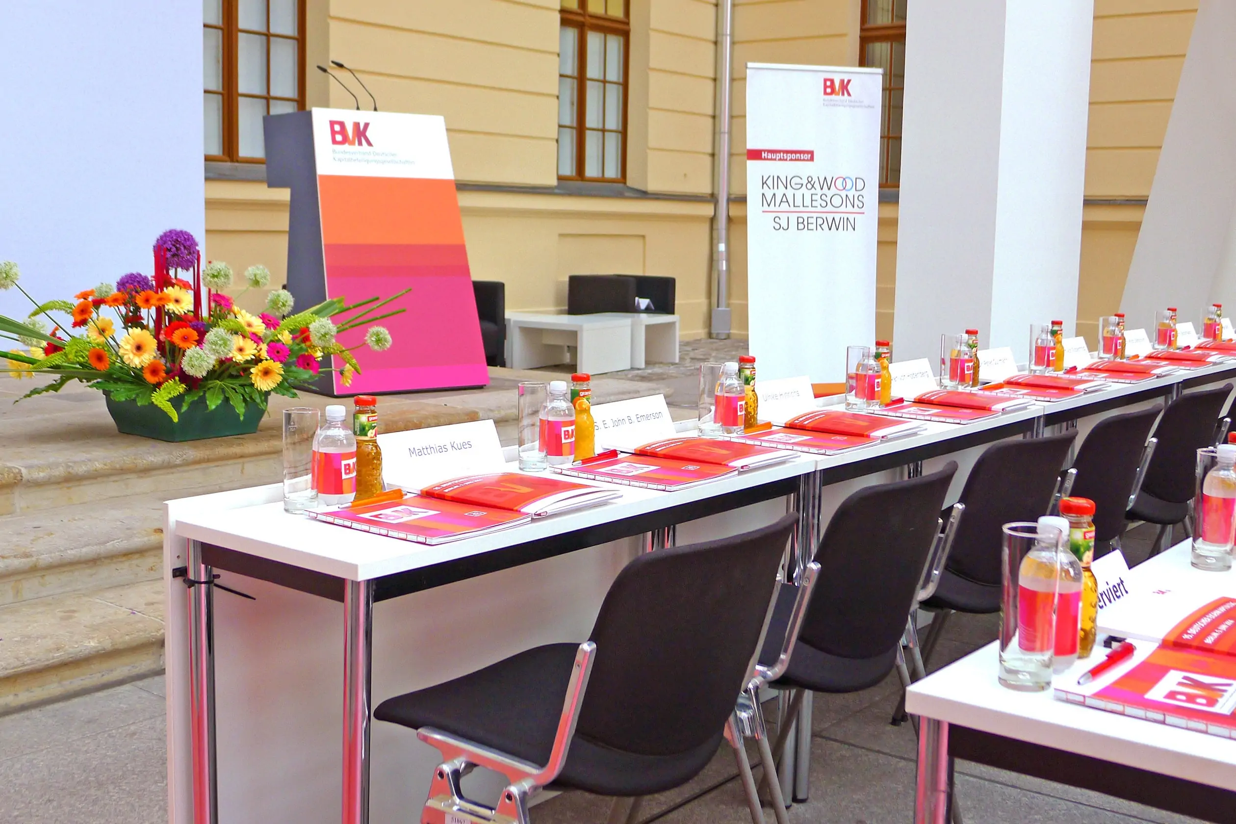 Tischreihen mit Drucksachen und Getränken für die Zuschauer des Events, gestaltet nach Richtlinien des Corporate Designs von BVK e.V. Im Hintergrund eine Blumendekoration und ein Pult, gehalten in der Farben von BVK e.V.