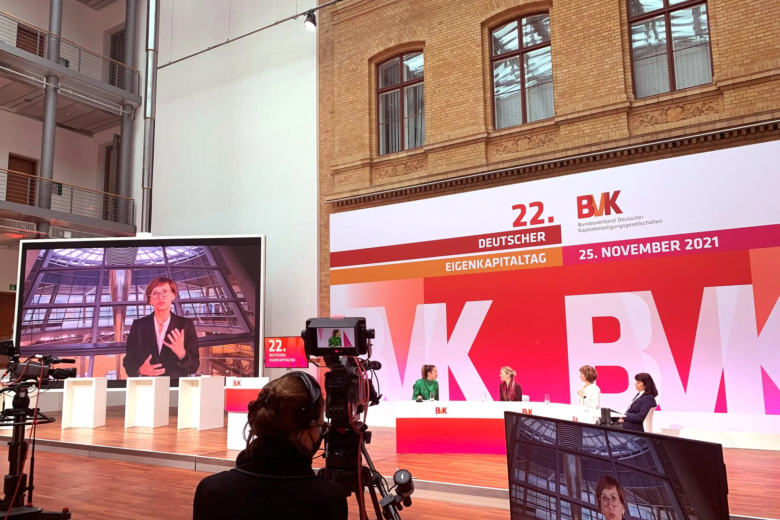 Im Vordergrund steht eine Frau hinter einer Kamera, die das Geschehen auf dem Podium des 22. Deutschen Eigenkapitaltags auf eine große Videoleinwand im Hintergrund überträgt. Auf dem Podium sitzen vier Personen hinter einem geschwungenem Pult und diskutieren miteinander. Dahinter befindet sich eine große Bühnenwand mit BVK-Branding.