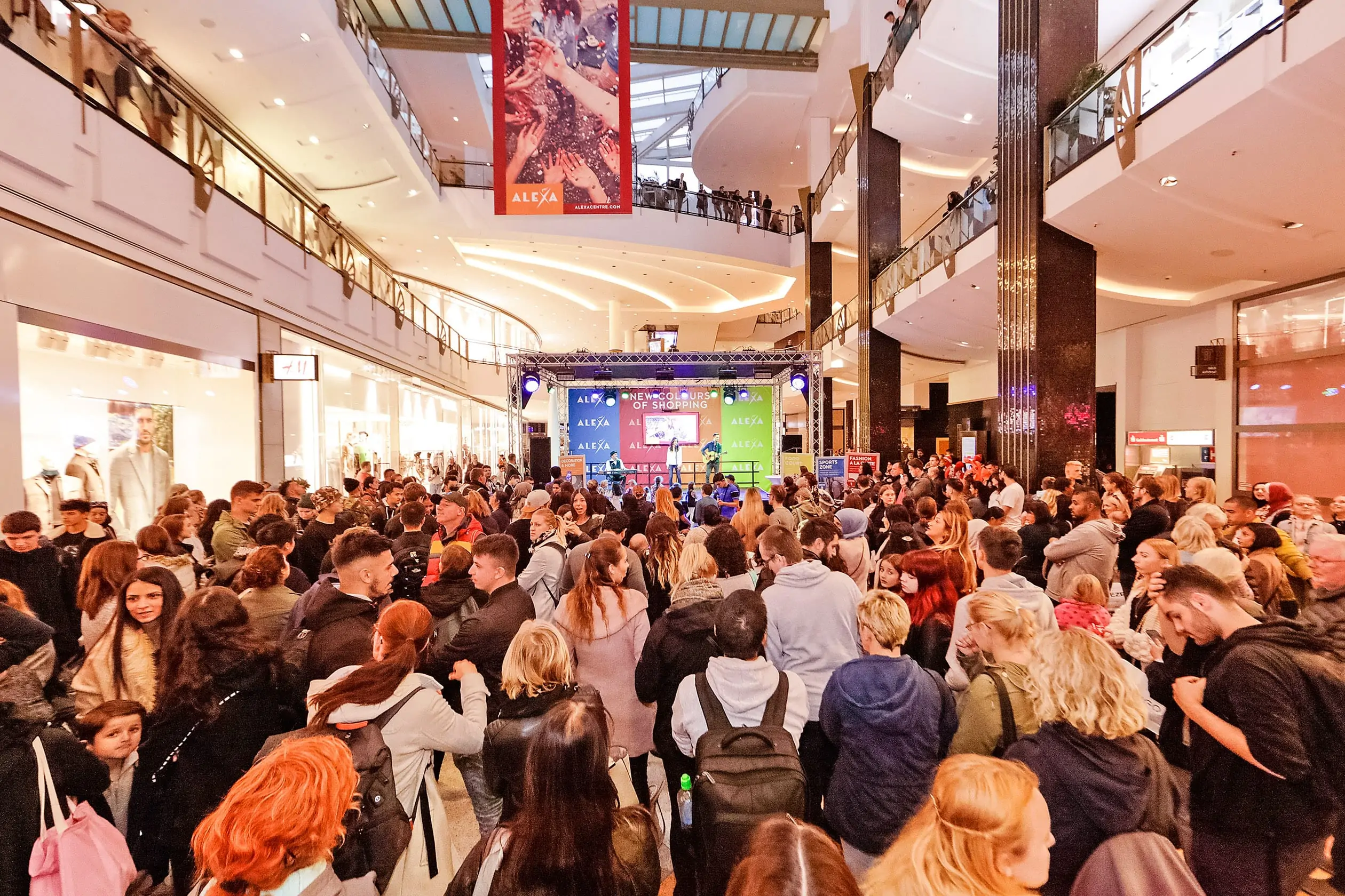 Im Metropolis Court des ALEXA Shoppingcenters findet eine Musikshow im Rahmen des Events ALEXA NEW COLOURS OF SHOPPING. Im Vordergrund sind zahlreiche Zuschauer zu sehen, die eine Musikshow verfolgen. Im Hintergrund steht die Bühne mit einer dreiköpfigen Musikband.