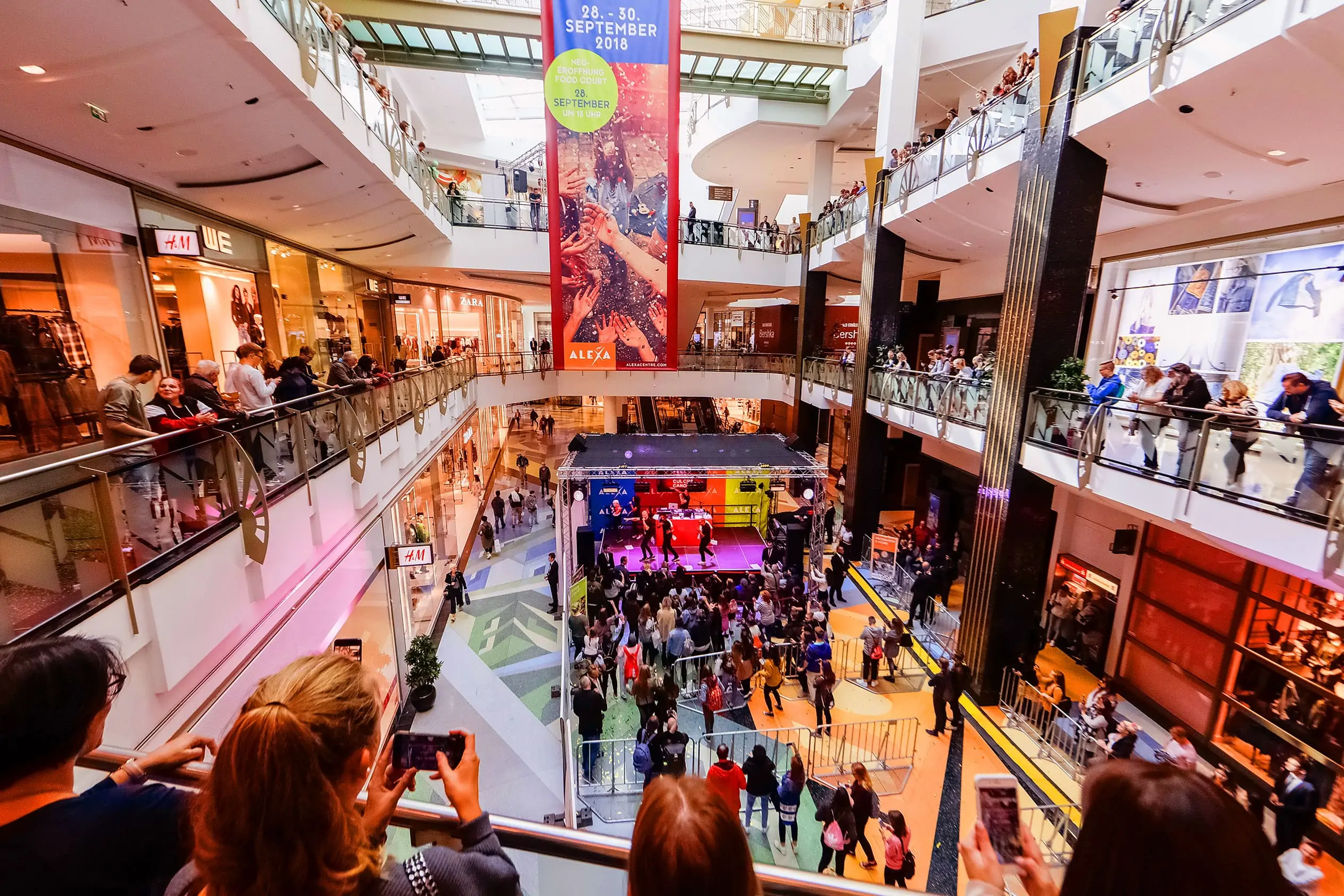 Eine prominente Musikband singt und tanzt auf der Bühne des Metropolis Court im ALEXA Shoppingcenter, im Rahmen des Events ALEXA NEW COLOURS OF SHOPPING. Vor der Bühne und auf den Rängen sind zahlreiche Zuschauer, die die Show verfolgen.