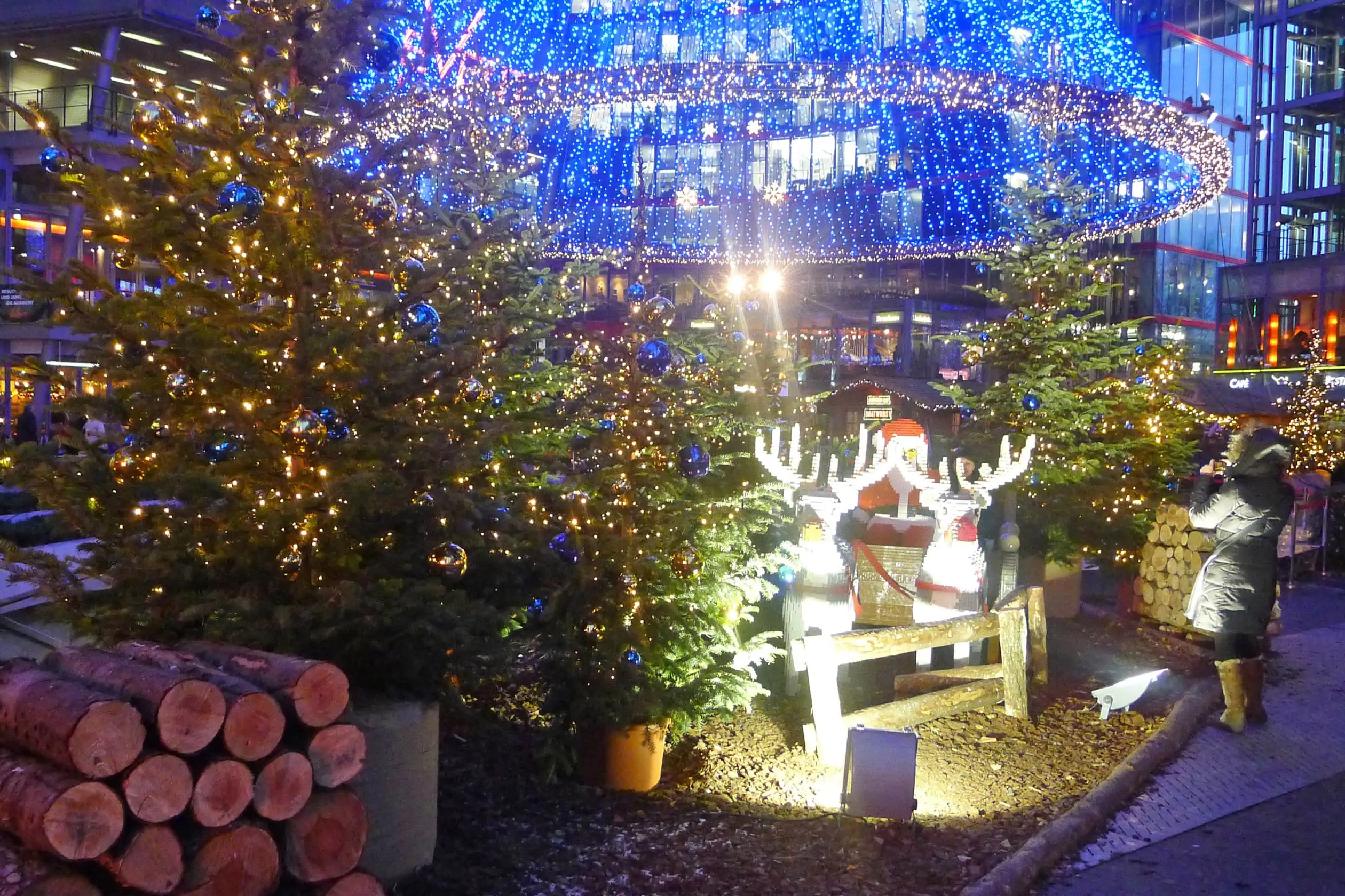 Ein Gespann aus zwei Rentieren und Weihnachtsmann, zusammengebaut aus Lego-Steinen, umgeben von weihnachtlich geschmückten Tannenbäumen. Im Hintergrund ein weihnachtlicher Trichter aus von oben herabhängender, blau leuchtender Lichterketten. SONY Center am Potsdamer Platz Weihnachtsaktionen Magic Christmas