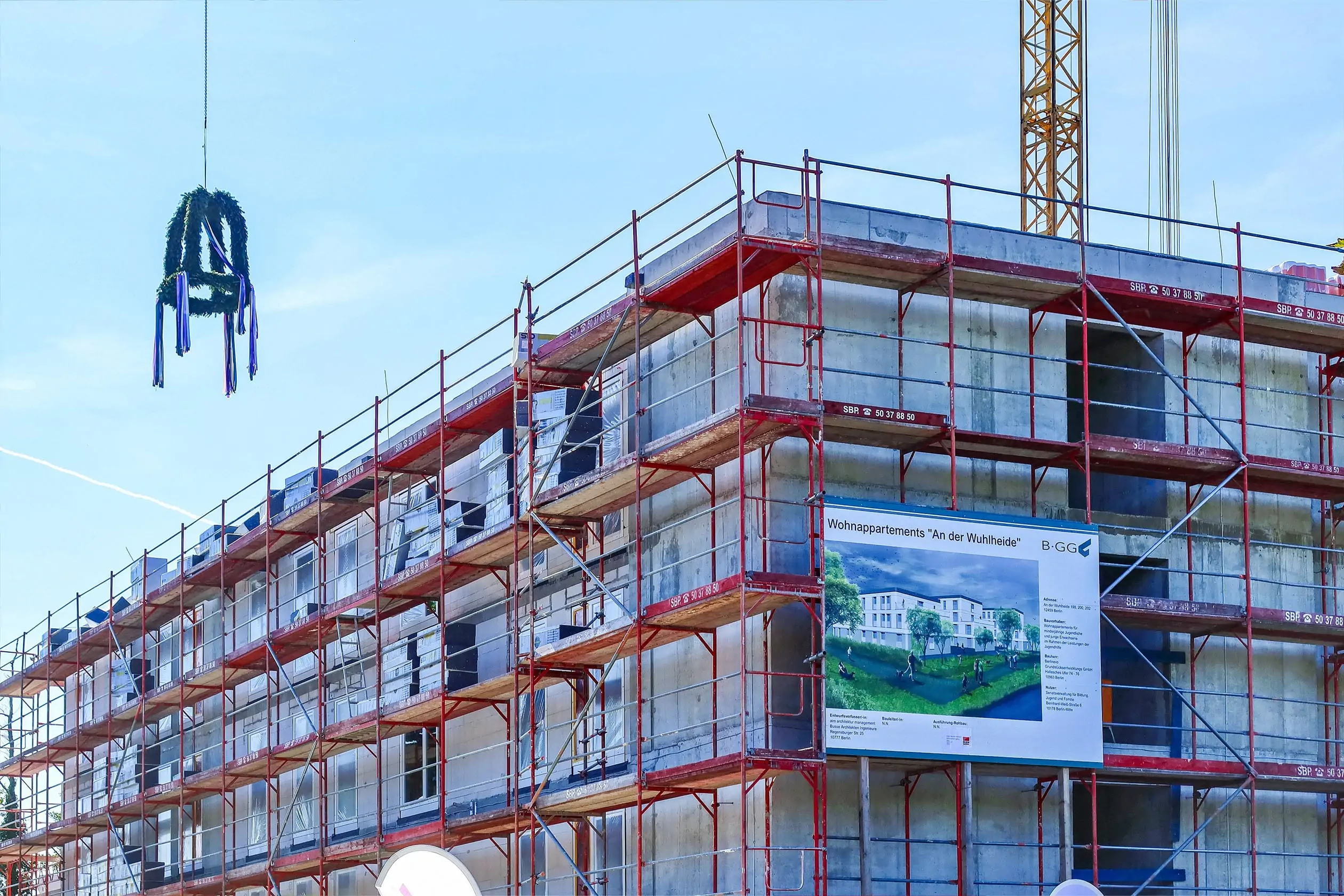 Ein Richtkranz schwebt über dem Bauprojekt von berlinovo in der Wuhlheide. Das Gebäude ist vom Baugerüst umhüllt. Ein Bauschild ist auf dem Baugerüst befestigt. Richtfest An der Wuhlheide.