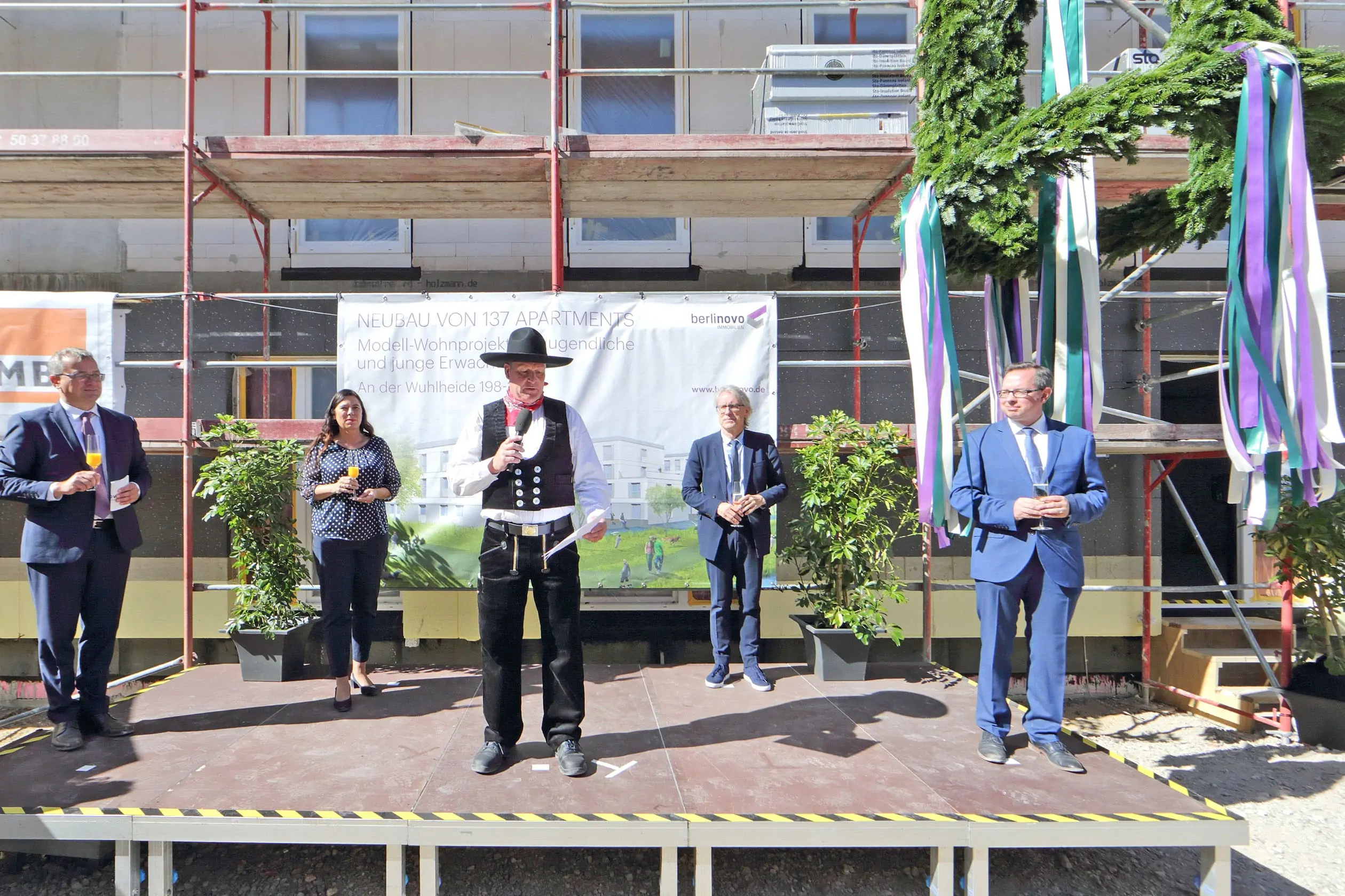 Fünf Personen, die als Redner auf dem Richtfest eines belinovo-Bauprojekts in der Wuhlheide auf einer Bühne vor dem Baugerüst stehen.