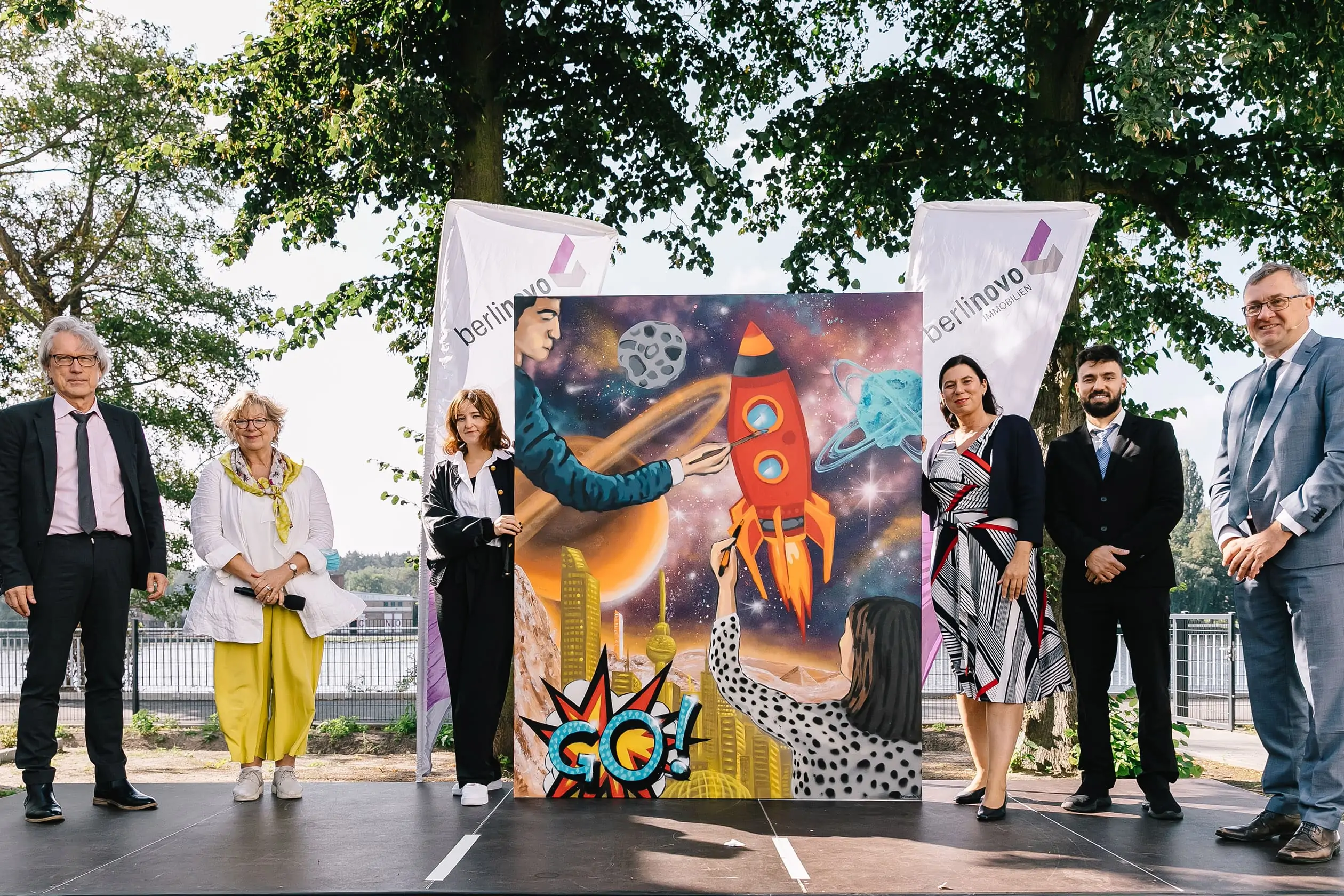 Sechs Personen auf der Bühne bei der Schlüsselübergabe eines Bauprojekts von berlinovo in der Wuhlheide. Sie präsentieren ein futuristisches Gemälde, das aus Anlass der Schlüsselübergabe angefertigt wurde.