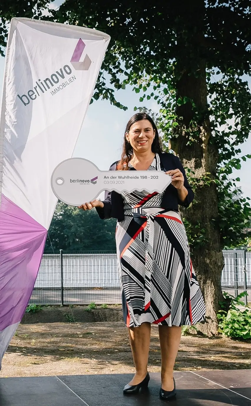 Eine Person auf der Bühne bei der Schlüsselübergabe eines Bauprojekts von berlinovo in der Wuhlheide. Sie präsentiert einen symbolischen Schlüssel, der aus Anlass der Schlüsselübergabe angefertigt wurde. Hinter den Personen sind Banner mit Branding von berlinovo.