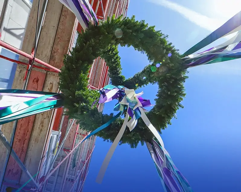 Auf dem Richtfest eines Bauprojekts von berlinovo in der Wuhlheide wird ein Richtkranz mit weissen, grünen und violetten Bändern, von unten Fotografiert, entlang eines Baugerüsts nach oben gezogen.