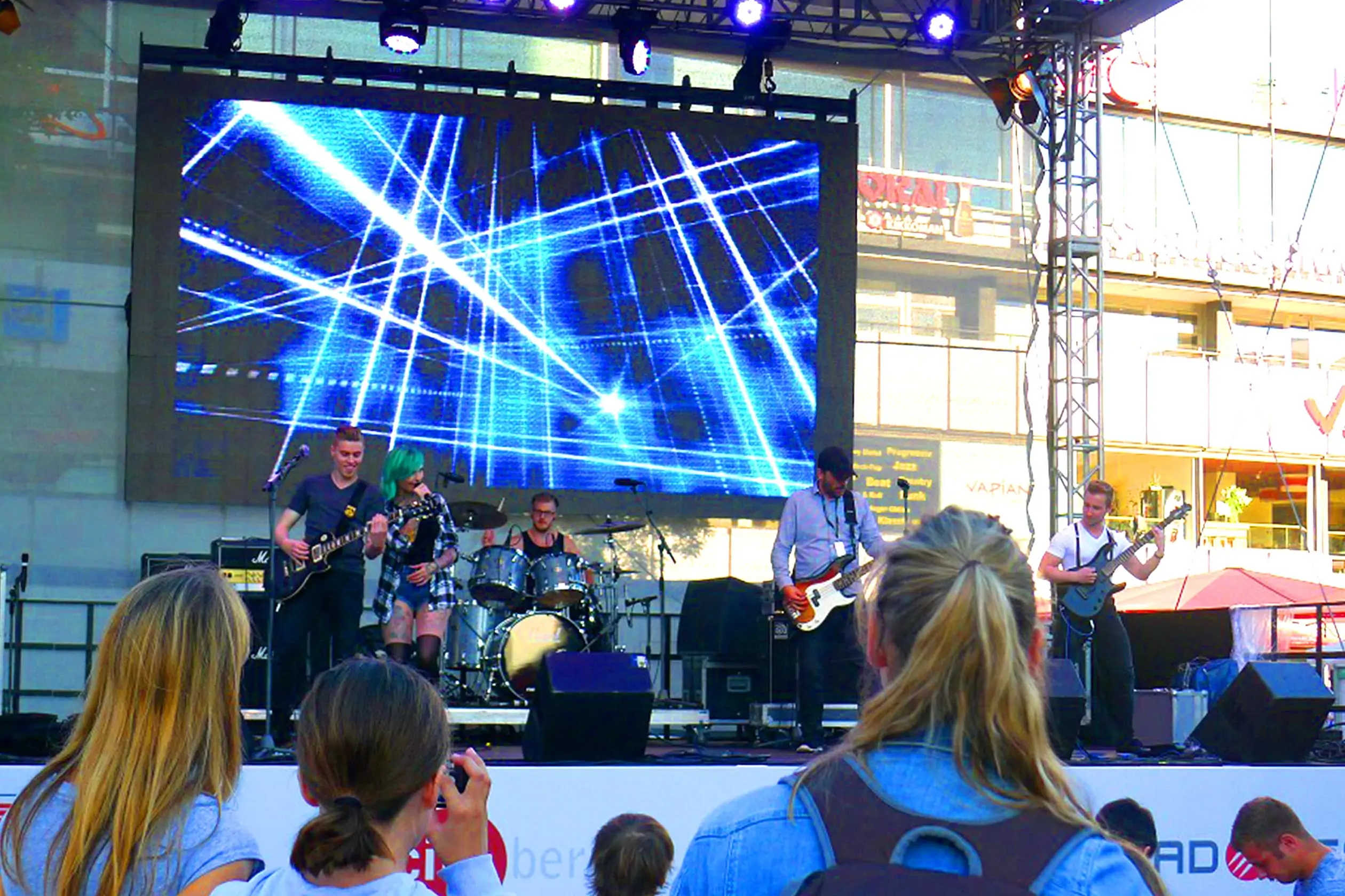 Eine Aufnahme eines der 30 Liveacts auf dem Breitscheidplatz in Berlin, auf dem das Event Berlin Celebrates IFA stattfindet. Auf der Bühne spielt eine Band. Von links Nachrechts sind zu sehen: ein Gitarrist, eine Sängerin, ein Schlagzeuger, ein Bassist und ein weiterer Gitarrist. Hinter der Band wird eine Lichtshow auf einer Leinwand vorgeführt. Vor der Bühne stehen Zuschauer. Trendshow Kurfürstendamm City West