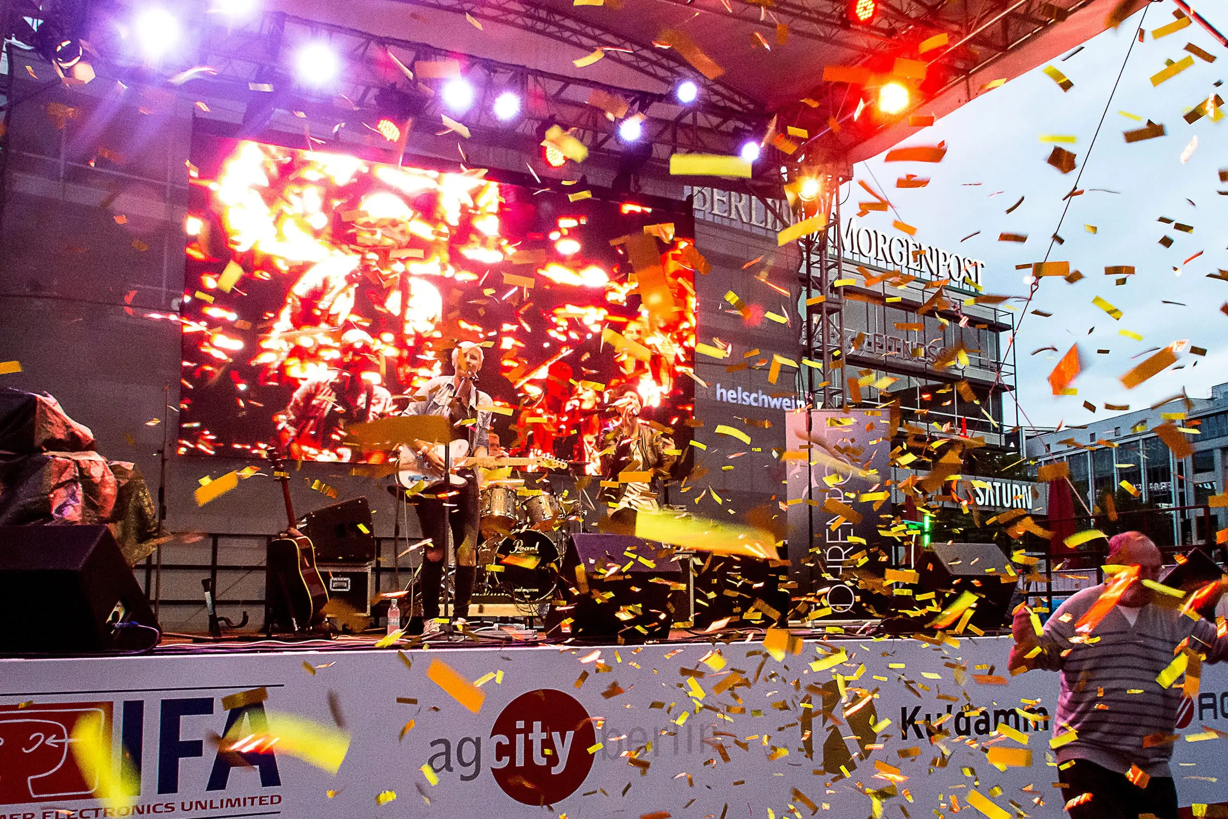 Eine Aufnahme eines der 30 Liveacts auf dem Breitscheidplatz in Berlin, auf dem das Event Berlin Celebrates IFA stattfindet. Auf der Bühne sind eine Sängerin mit Gitarre, und dahinter ein Schlagzeuger währen eines Konzerts zu sehen. Ein Konfetti-Regen aus goldenen Papierstreifen fliegt durch die Luft. Kurfürstendamm City West