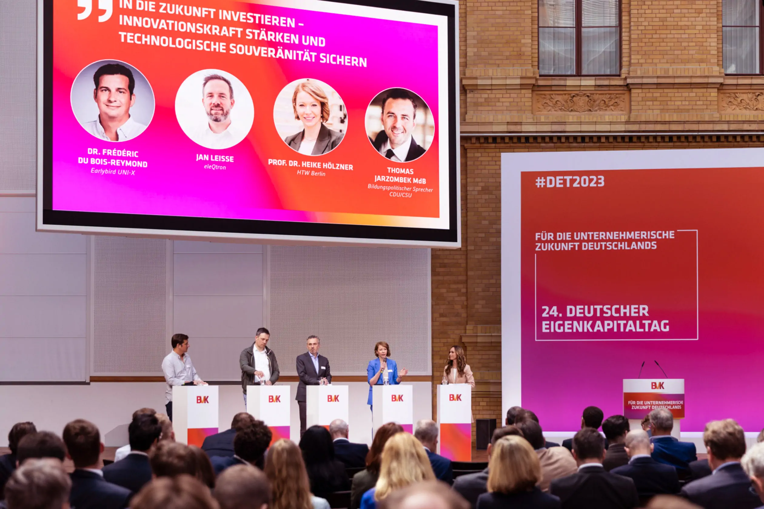 Auf dem Podium des 24. Deutschen Eigenkapitaltags führen fünf Referenten, hinter Rednerpulten mit BVK-Branding stehend, eine Podiumsdiskussion durch. Hinter den Referenten und rechts daneben ist eine große Videoleinwand und eine große Bühnenwand mit BVK-Branding zu sehen. Vor den Referenten sind mehrere Zuschauerreihen zu sehen.