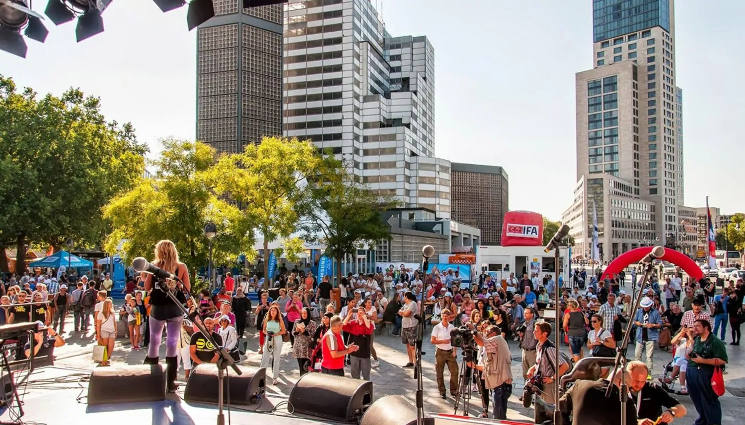 Eine Aufnahme auf dem Breitscheidplatz in Berlin, auf dem das Event Berlin Celebrates IFA stattfindet. Im Vordergrund ist eine Bühne zu sehen. Ein Musikerin spielt vor zahlreichem Publikum, einem Kameramann und mehreren Fotografen. Im Hintergrund sind Hochhäuser zu sehen.