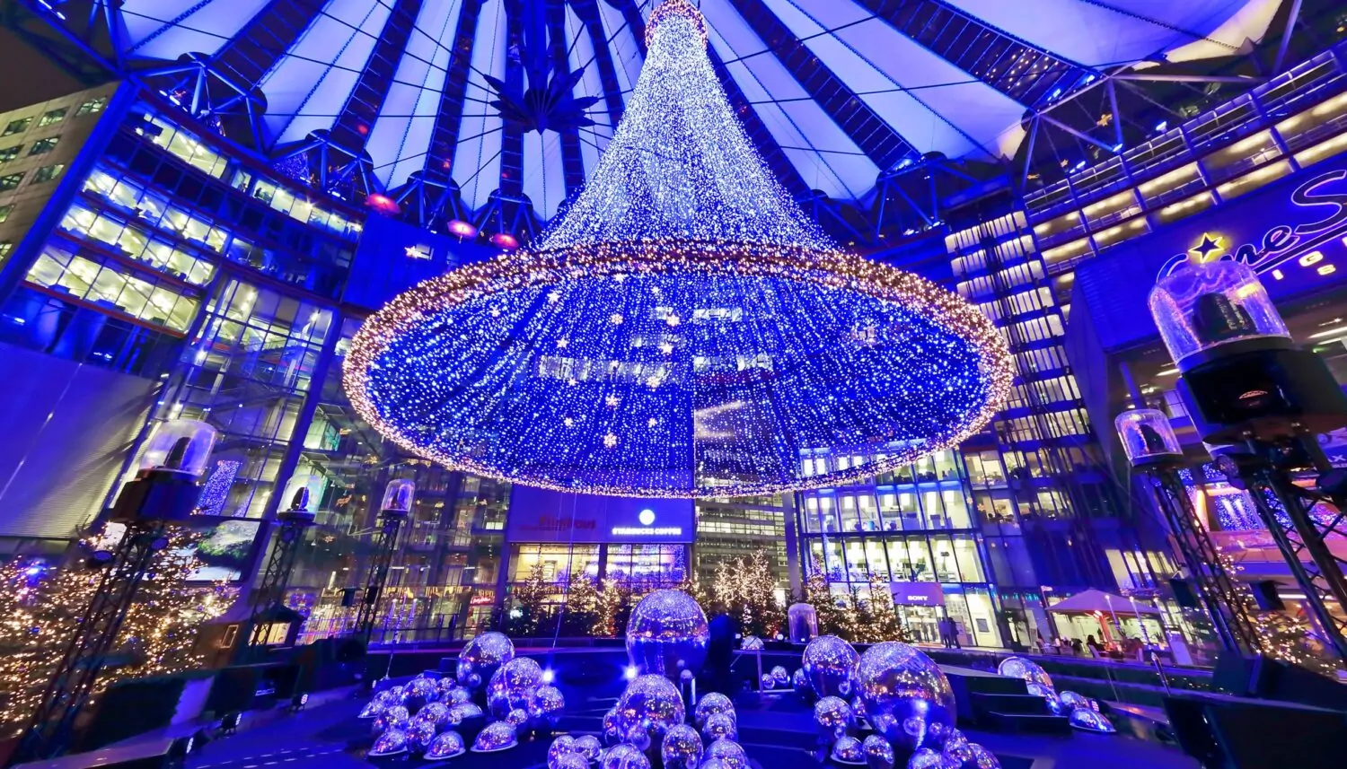 Weihnachtsdekoration des Sony Centers aus einem Trichter aus von oben herabhängender, blau leuchtender Lichterketten, großen, blau leuchtenden Kugeln, Tannenbäumen mit Lichterketten und Weihnachtsbeleuchtung. Der Innenraum des Sony Centers und das Dach ist in blaues Licht getaucht.