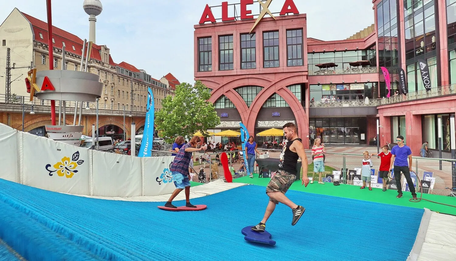 Für das Event ALEXA – THE WAVE wurde vor dem Eingang zum ALEXA Shoppingcenter eine künstliche Surfing-Welle aufgebaut. Im Vordergrund sind zwei junge Männer zu sehen, die auf Surfbrettern die Welle reiten. Im Hintergrund ist die Moderatorin des Events und wartende Besucher auf dem Ravelinplatz vor dem Eingang zum ALEXA Berlin zu sehen.