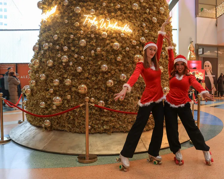 Zwei Frauen in roten Weihnachtskostümen und Rollschuhen posieren vor einem riesigen, gold‑verzierten Weihnachtsbaum in der ALEXA‑Mall in Berlin.